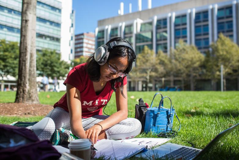 Student wearing headphones and an MIT t-shirt sits on the grass and writes in a notebook.