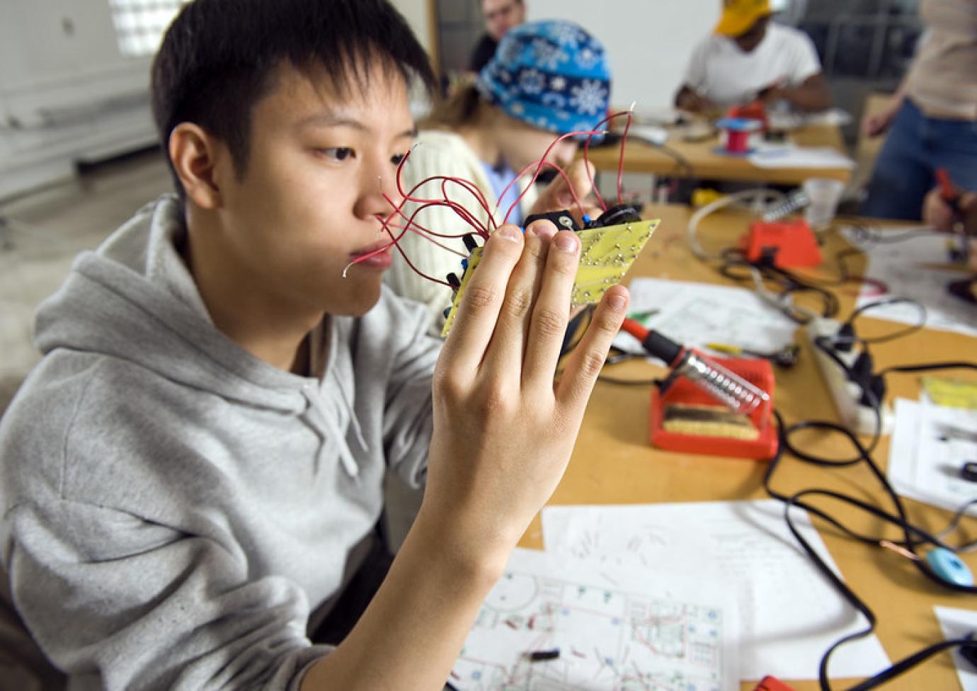 A student holds up a circuit board while other students work in the background.