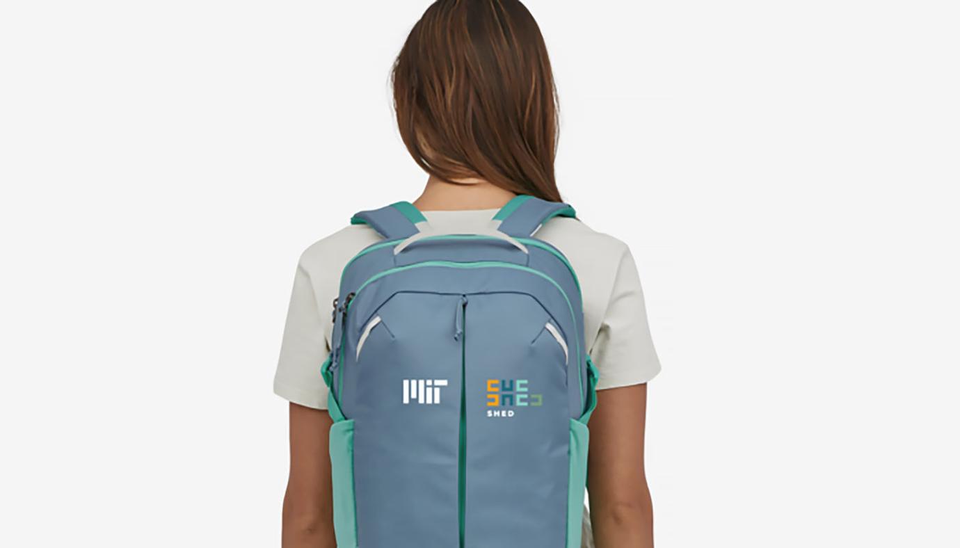 Person wearing a backpack with a white MIT logo on the left side and the SHED logo on the right side.
